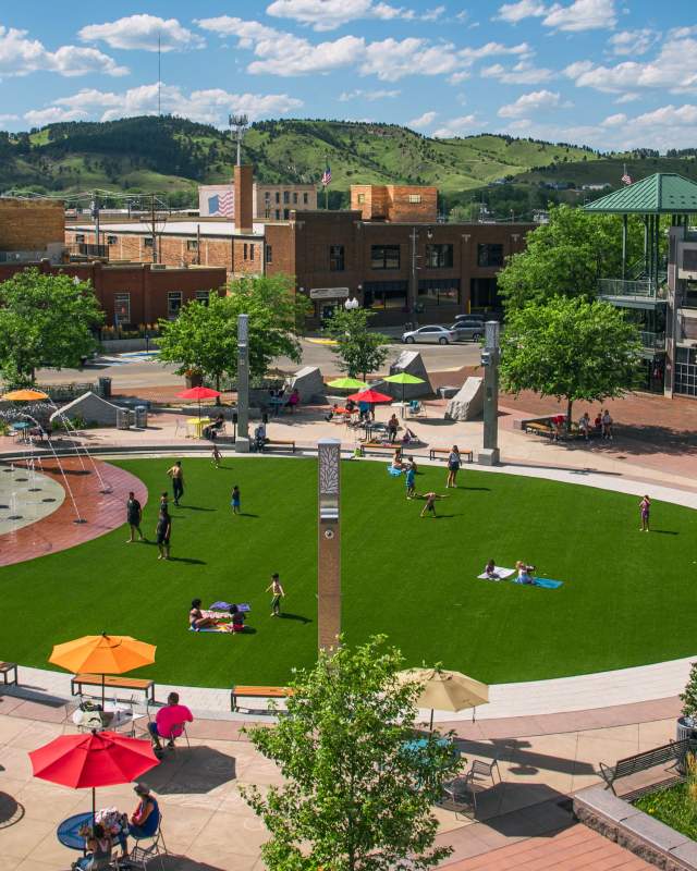 Main Street Square with lush green grass, people relaxing, kids playing near fountains, umbrellas providing shade, and historic brick buildings surrounding.