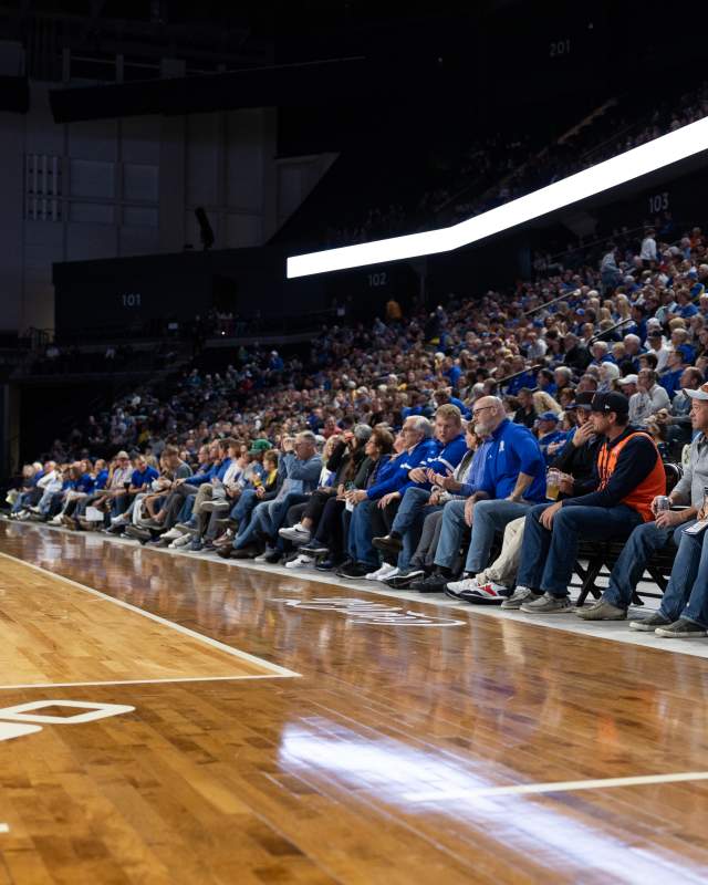 Large crowd seated courtside and in arena stands during a basketball game, with fans watching the action from rows along the hardwood floor under bright indoor lighting.