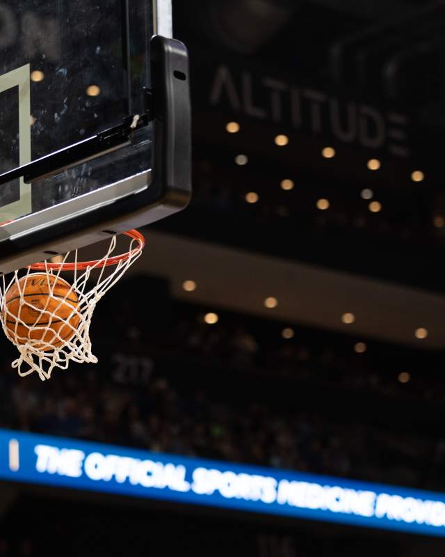 A basketball swishes through a hoop in a brightly lit arena. Mood lighting from arena suites are visible in the background, but no fans are clearly seen.