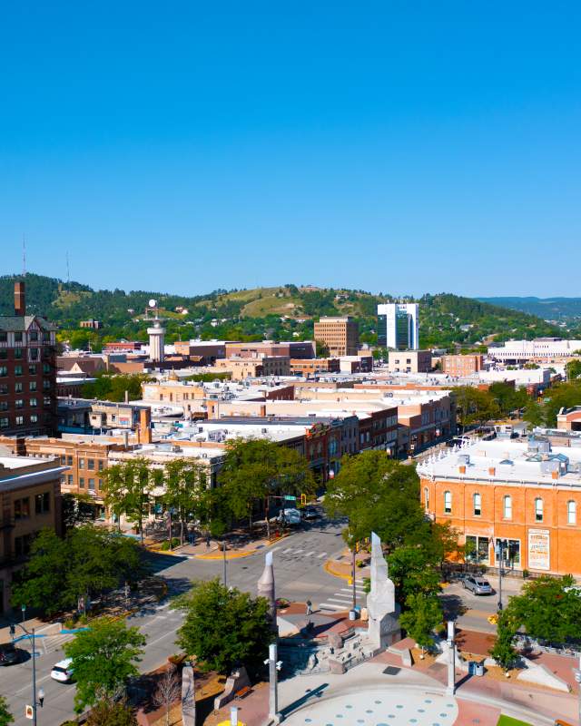 Aerial view of downtown Rapid City featuring historic brick buildings, tree-lined streets, and the Hotel Alex Johnson sign, with green Black Hills in the background under a clear blue sky.
