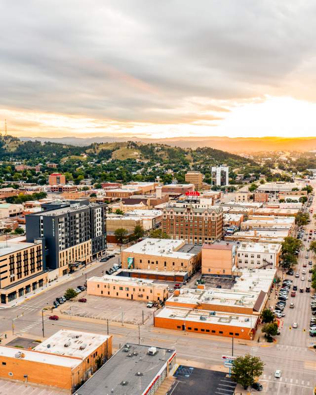 Aerial view of downtown Rapid City at sunset, with downtown streets lined by low-rise buildings, cars, and parking lots. Rolling green hills surround the city, while dramatic clouds stretch across the sky and golden sunlight glows on the horizon.
