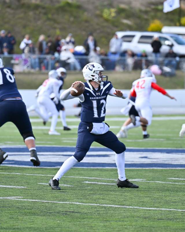 The quarterback for South Dakota Mines, wearing a navy blue uniform with a white #12 on the front, throws a pass in the foreground as players from both teams are visible in the background.
