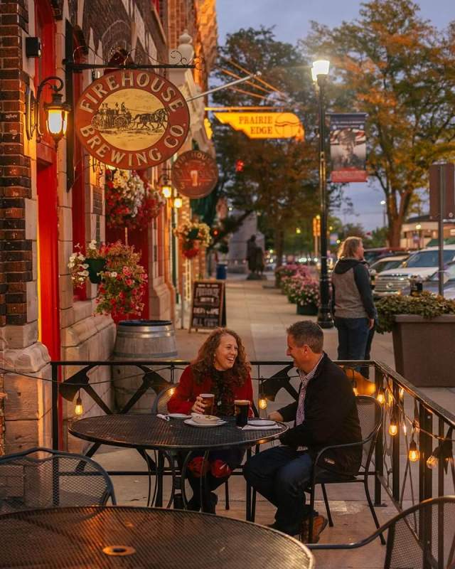 A couple enjoys an evening on an outdoor patio at Firehouse Brewing Co. with brick walls and warm lighting, surrounded by flowerpots and streetlamps on a bustling city street.