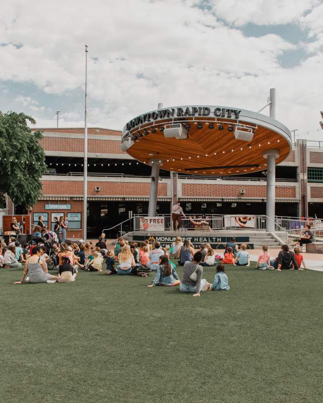A crowd of adults and children sits on a grassy plaza facing an outdoor stage beneath a circular canopy labeled “Downtown Rapid City.” The stage is set in a downtown square surrounded by brick buildings, string lights, and a parking structure. People listen to a presenter on stage under a partly cloudy sky, creating a lively community gathering atmosphere.