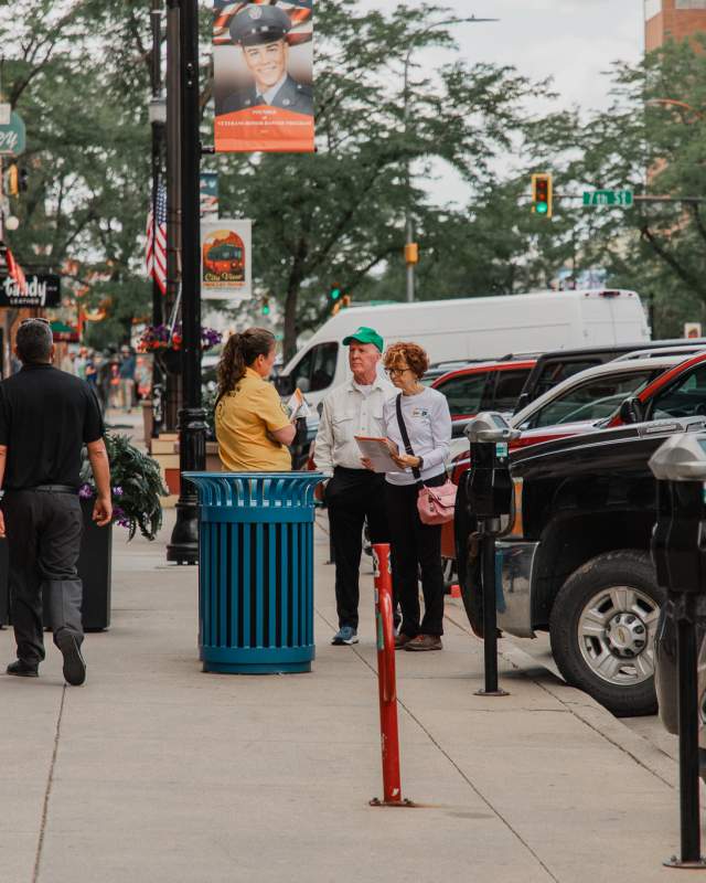 Downtown Rapid City Ambassador in a yellow shirt speaks with visitors on a busy sidewalk lined with storefronts, signs, parked cars, and street banners.