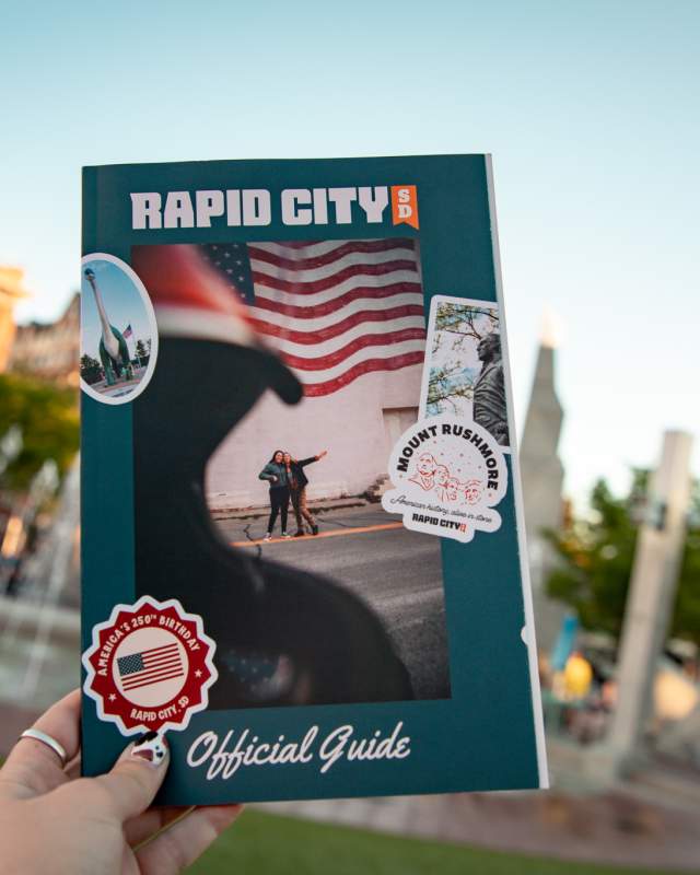 The cover of the Visit Rapid City guide is held up by a woman against the backdrop of the fountains at Main Street Square.