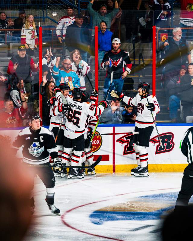 Hockey players in white jerseys gather for a group hug after scoring a goal. Fans behind the boards stand and celebrate with the team.