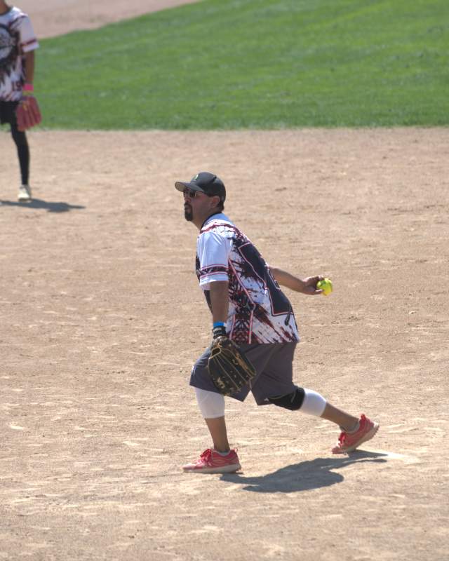 A softball pitcher in a patterned jersey and cap winds up to throw a bright yellow softball on a sunlit dirt field, with another player standing in the outfield behind him.