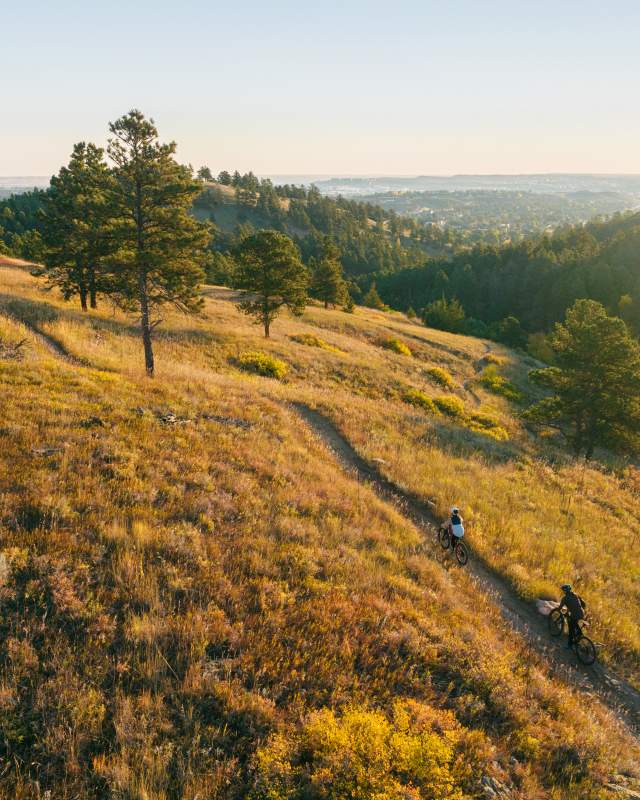 From a higher altitude, two bikers ride a trail along Skyline Drive in Rapid City in the early morning sunlight in a clear sky that casts a golden hue across the landscape.