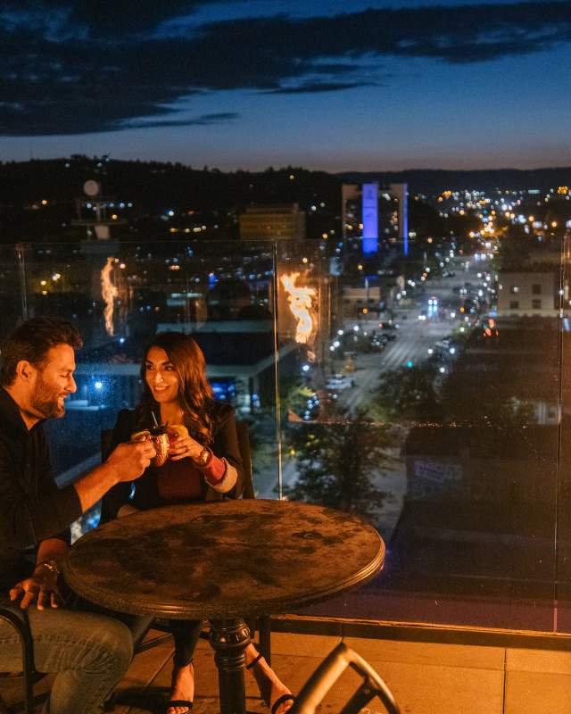 Couple enjoying drinks at a rooftop bar beside a glass fire feature, overlooking the glowing city lights of Rapid City at dusk. The pair sits at a small table, toasting mugs while the skyline and Black Hills foothills stretch into the evening background.