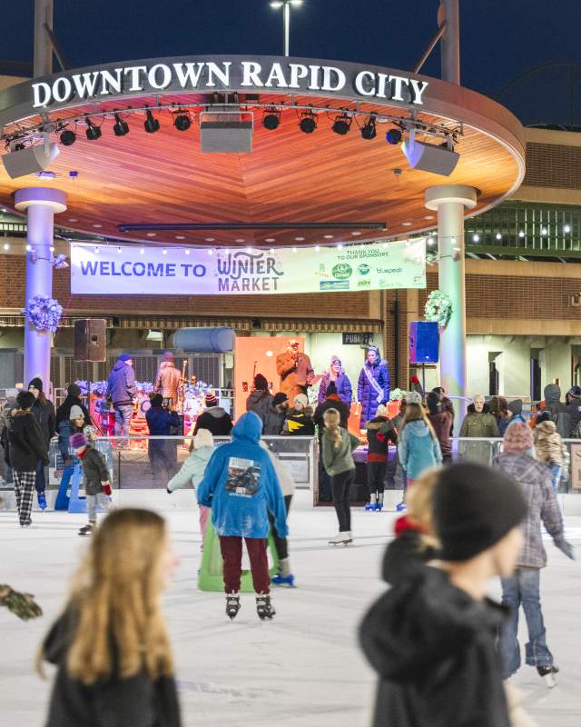 People skate on an ice rink in a lively winter market scene at night. A band performs on stage under a “Downtown Rapid City” sign, creating a festive atmosphere.