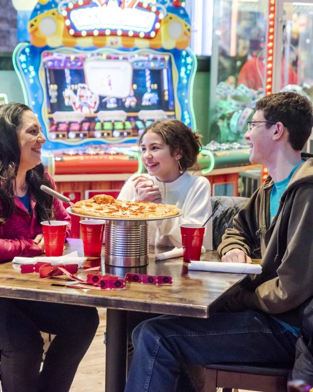 Family of three sitting around a table eating pizza with arcade games in the background at Press Start in Rapid City, SD