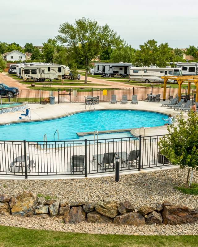 Outdoor pool with blue water heavily distorted by a digital glitch, partially revealing a drained pool basin with tiled walls, concrete edges, and a rocky border, with RVs and trees in the background under a clear sky.
