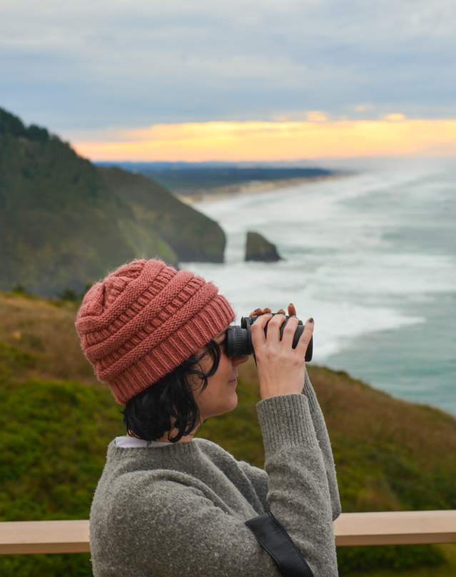 A person looks through binoculars at the ocean with coastal cliffs in the background