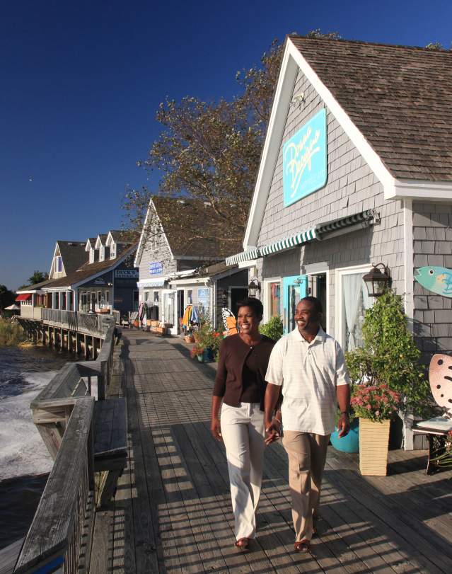 A couple strolls hand-in-hand along a scenic boardwalk by the water, surrounded by colorful shops and vibrant coastal flora.
