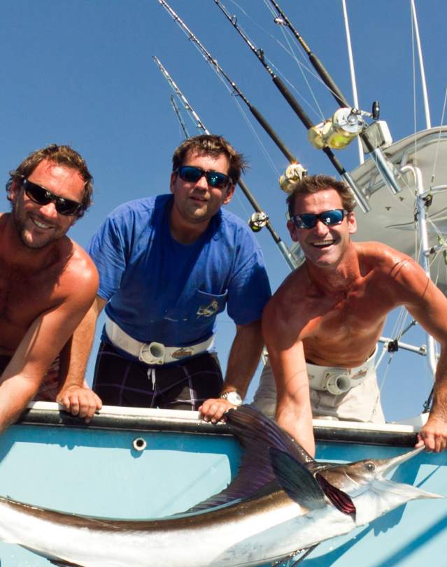 Three men on a fishing boat hold up a large fish, showcasing their catch under a clear blue sky.