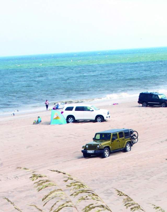 Off-road vehicles parked on Outer Banks sandy beach with visitors walking along the shore and turquoise water in bright sunlight.