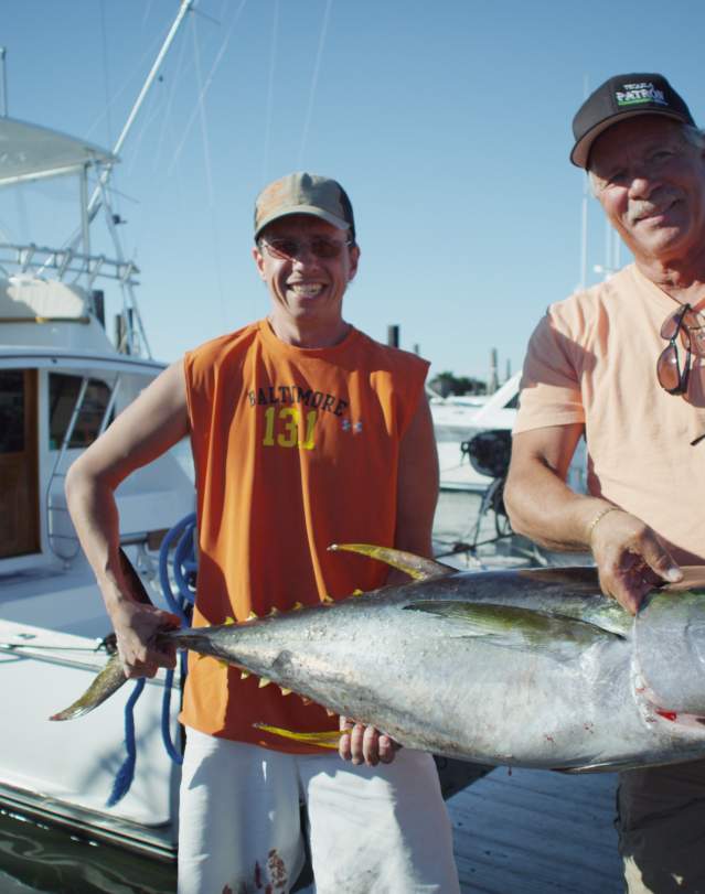 Two men proudly display a large fish beside a docked white boat under a clear blue sky.