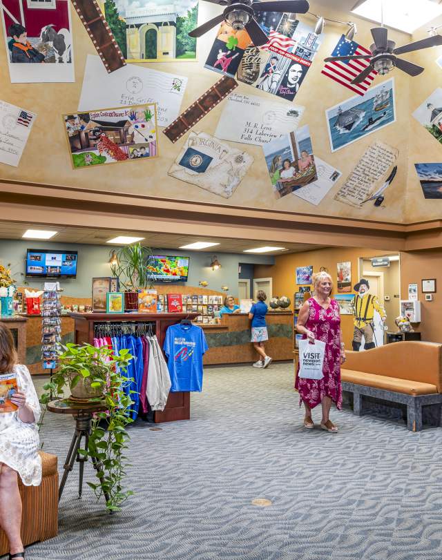 A woman is sitting reading the visitor guide magazine while another woman shops in a vaulted room with an artistic ceiling covered in places around Newport News