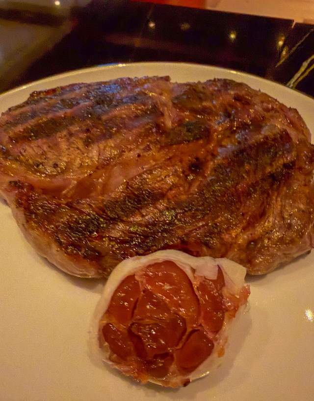 Dimly lit photo of a large steak with roasted garlic on the side