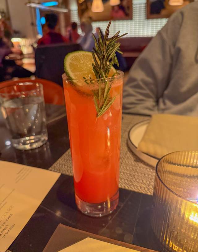 A tall glass with filled with a bold red mezcal cocktail, topped with a sprig of rosemary and large lime slice.