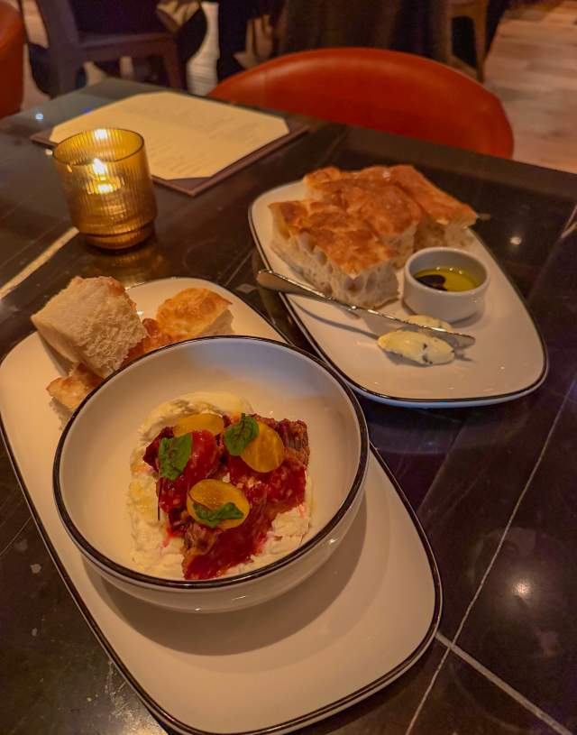 Two plates in a dimly lit restaurant, one offers a bowl of burrata and the second bread and butter.