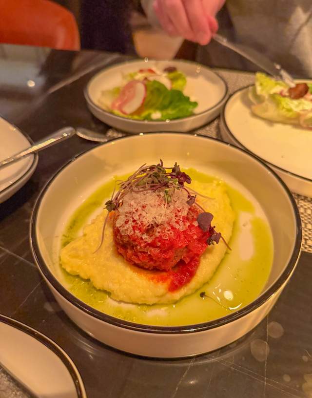 A dimly lit, slightly blurry iphone photo of a bowl containing a meatball on a bed of what appears to be polenta.