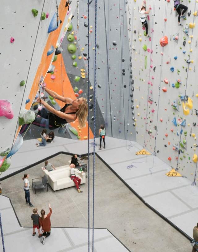 A photo looking down on the indoor climbing gym of CLIMB Bentonville. Lead ropes dangle in the foreground, with a climber on a rope contemplating their next move up the wall.