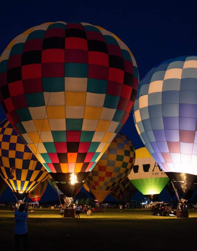 Colorful hot air balloons illuminated at night, set against a dark sky. Spectators gather around, creating a vibrant atmosphere.