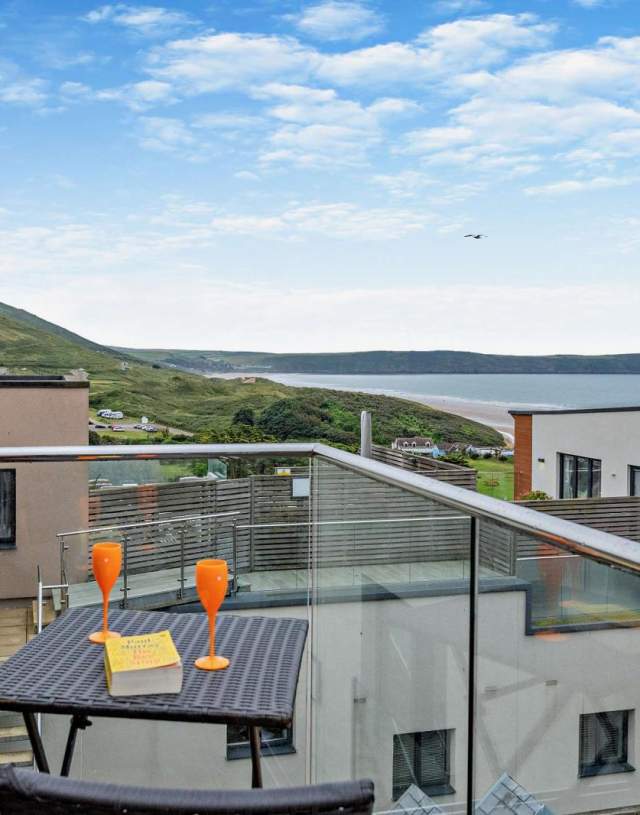 A modern balcony with a wicker table and chairs overlooks a coastal landscape. Two orange glasses and a book are on the table, creating a serene atmosphere at Byron's