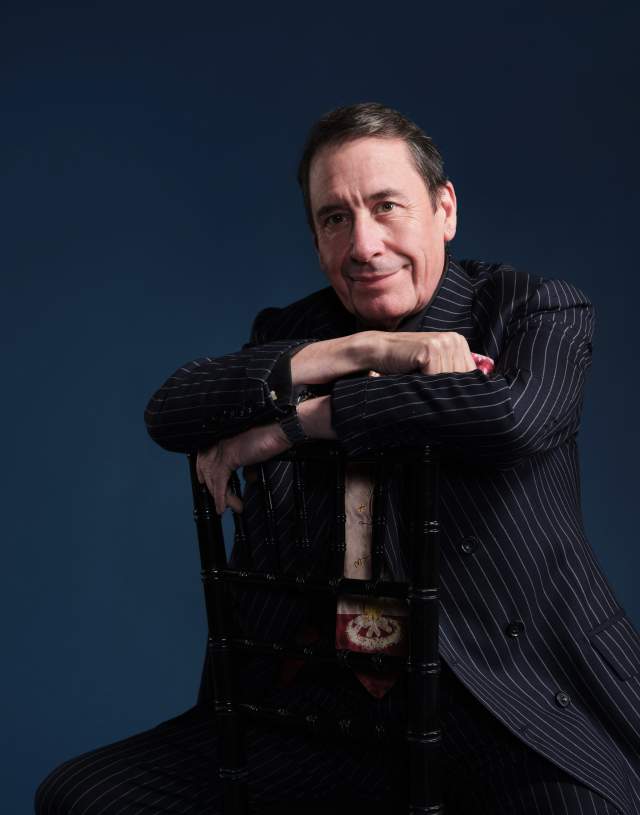 A man in a pinstripe suit leans on a chair, smiling warmly. The background is a solid dark blue, creating a professional and relaxed tone.