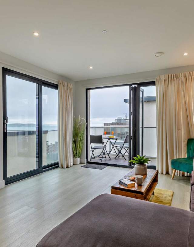 Modern living room with large windows, grey sofa, wooden coffee table with plants, and a balcony view. Light, airy ambience with neutral tones at Byron's