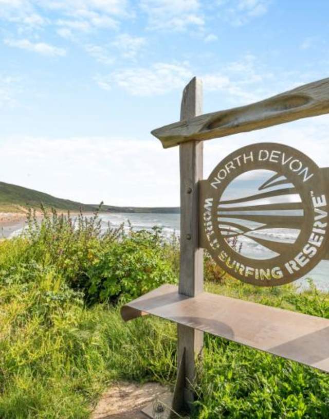 Wooden sign reads "North Devon World Surfing Reserve" with coastal landscape and ocean waves in the background under a blue sky.
