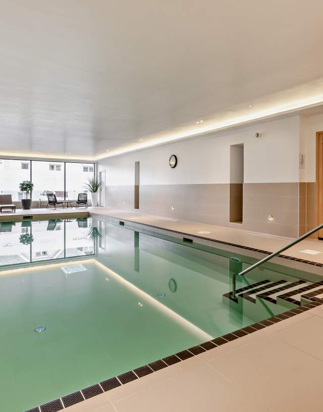 Luxurious indoor pool area with calm water reflecting soft lighting. Surroundings are beige tiles, lounge chairs, potted plants, and a wooden sauna at  Byron's