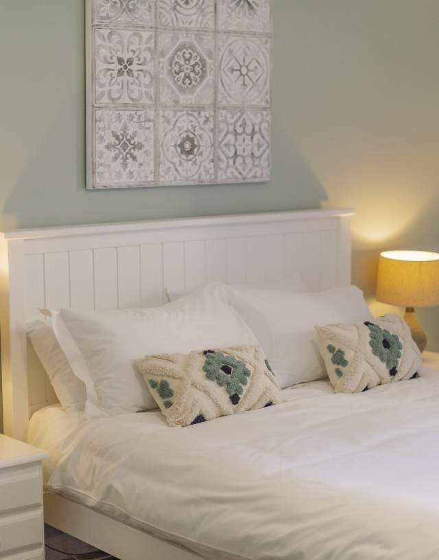 A cosy bedroom with a white bed, soft pillows, and patterned cushions. Warm lighting from two lamps creates a calm, inviting atmosphere. A decorative tile artwork hangs above the bed.