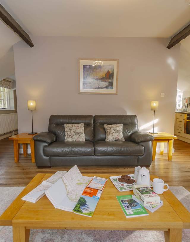 Cosy living room with a leather sofa, armchair, and wooden coffee table. Maps, brochures, and a teapot on the table. Warm, inviting atmosphere.