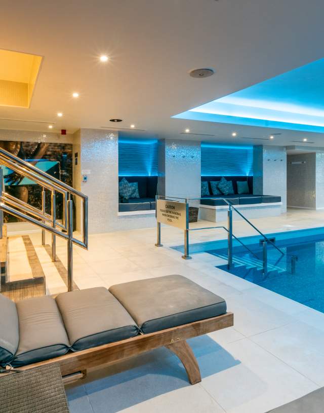 Indoor pool area with modern design features, such as a blue-lit ceiling and sleek lighting. There are cushioned lounge chairs and glass doors leading outside, creating a serene ambience.