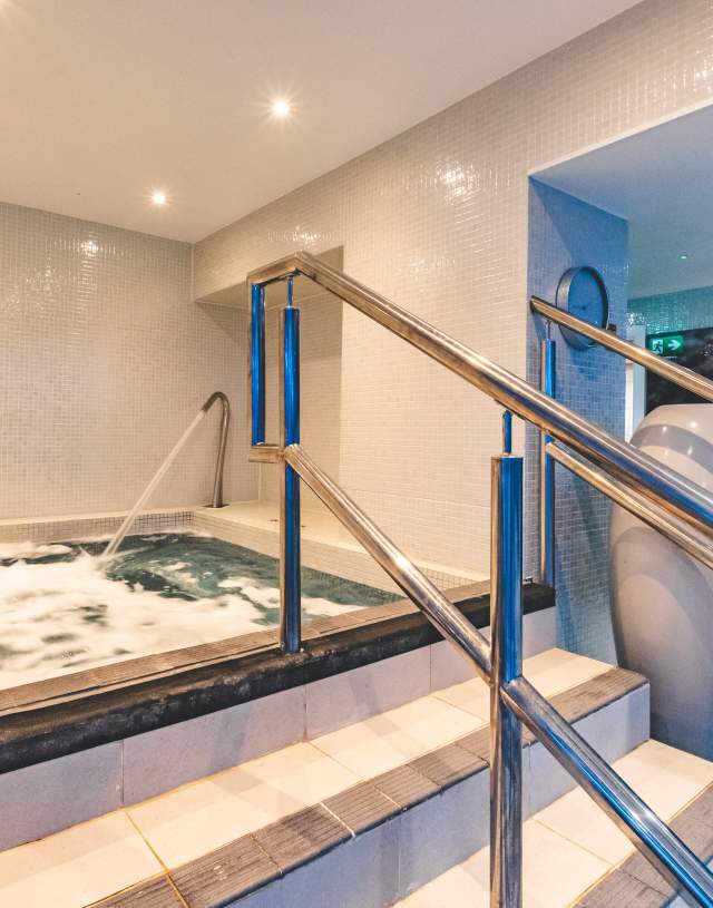 Indoor spa room with a bubbling jacuzzi, stainless steel handrails, and tiled walls. Soft lighting creates a relaxing, tranquil ambiance.