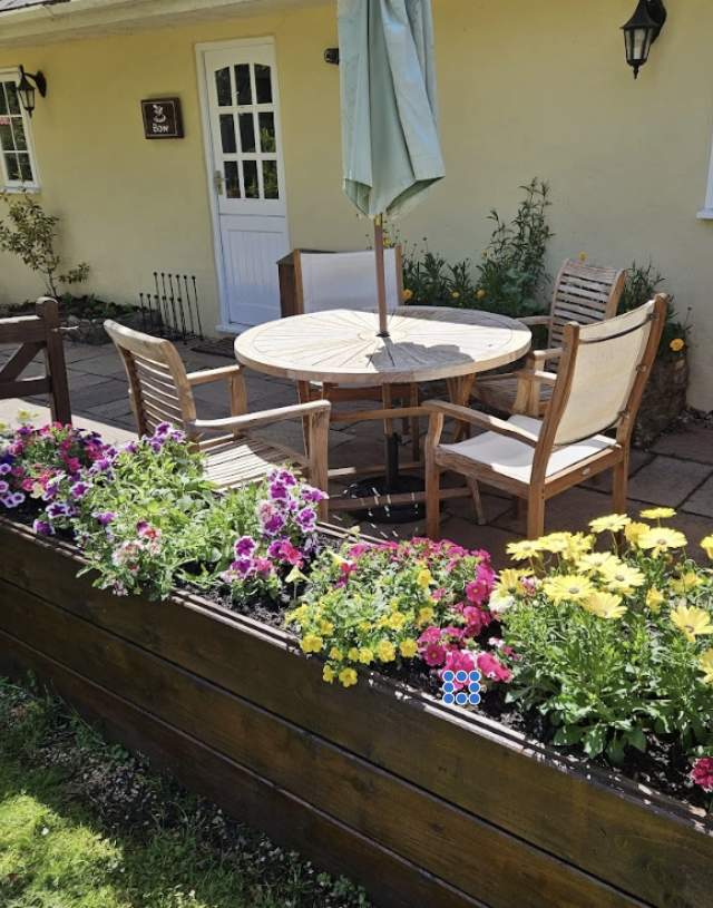 A cosy patio with wooden chairs and a round table shaded by a green umbrella. Vibrant yellow and pink flowers in a raised bed, backed by a yellow cottage.