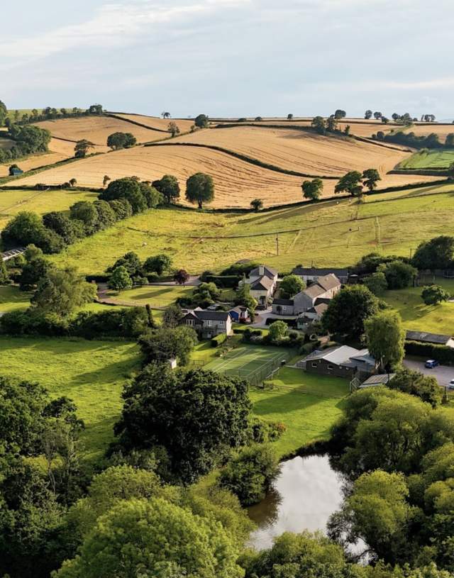 Rolling countryside with a patchwork of green and golden fields, a small village of houses, and lush trees surrounding a reflective pond, under a clear sky.