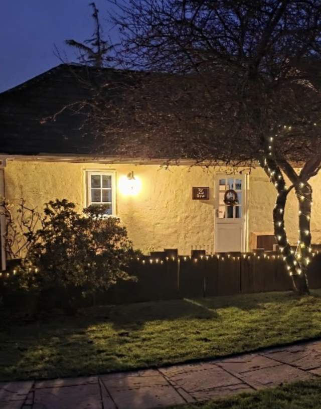 A cosy, warmly lit cottage at dusk with glowing fairy lights draped on trees and bushes. A path leads to the door, creating an inviting, tranquil scene.