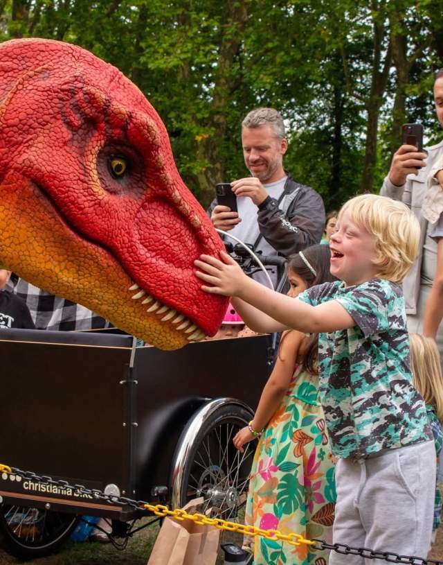 A young boy joyfully touches a bright red and orange dinosaur puppet, surrounded by excited onlookers taking photos in a lively outdoor setting.