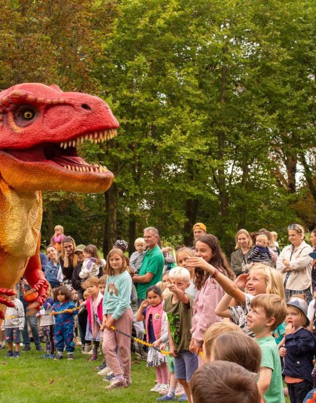 A vibrant red and orange dinosaur costume entertains an excited group of children and adults in a lush park, creating a fun and playful atmosphere.