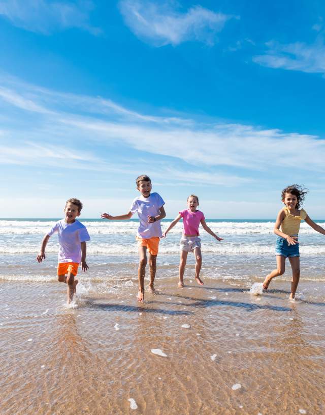 Four children joyfully run through shallow waves on a sunny beach, under a clear blue sky. Their laughter and energy evoke feelings of happiness and freedom.