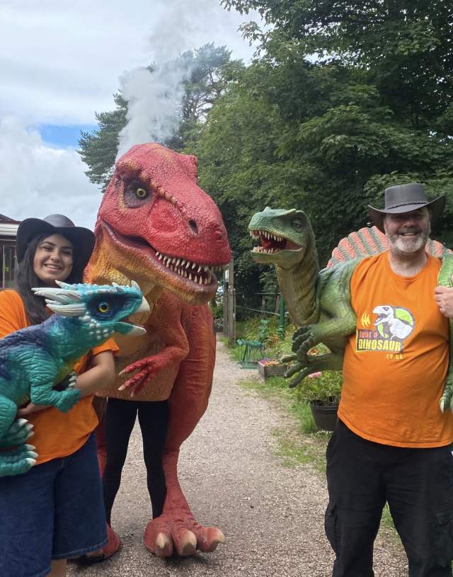 Two people in orange shirts and hats hold colorful dinosaur toys. A person in a dinosaur costume stands in between. There's a red train and trees nearby.