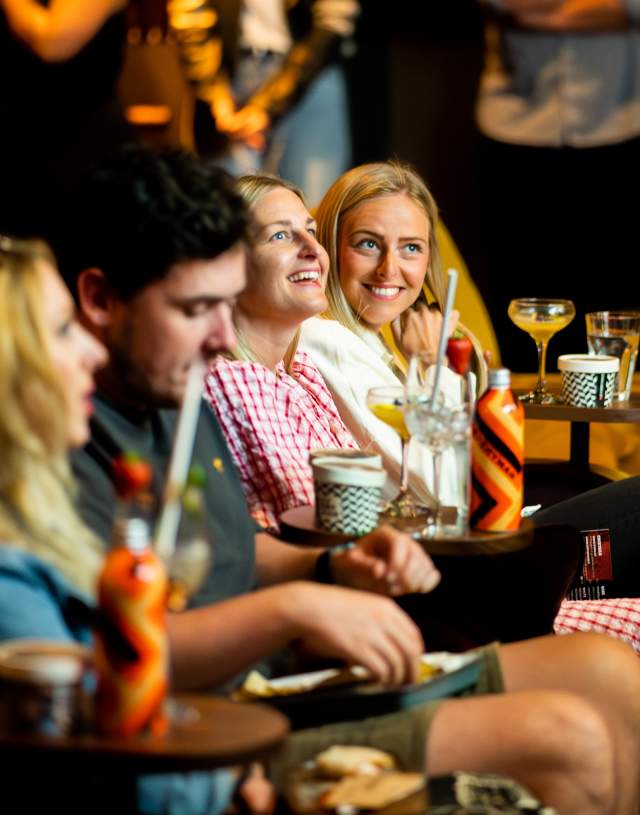 A group of friends in a cosy setting share laughter and drinks. They sit on plush seats with snacks and beverages on small tables, creating a warm, joyful atmosphere.