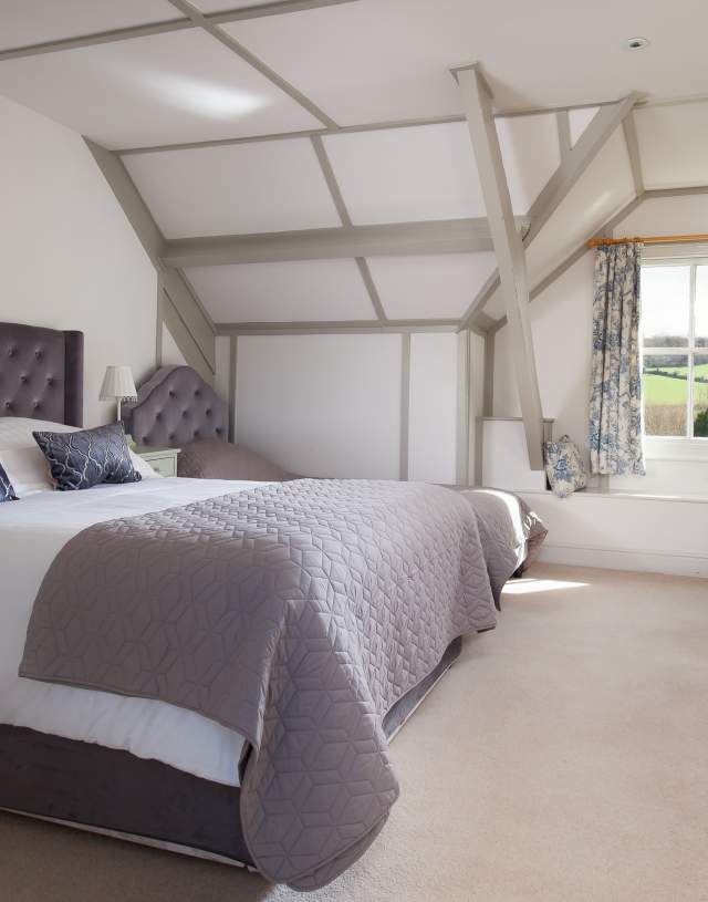 Spacious bedroom with a plush, tufted headboard, soft gray bedding, and elegant decor. A window with scenic countryside views and floral curtains adds charm.