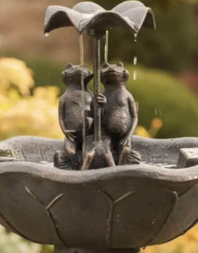 A stone fountain with two frog sculptures holding an umbrella, surrounded by lush garden foliage. It's a serene and whimsical garden scene.
