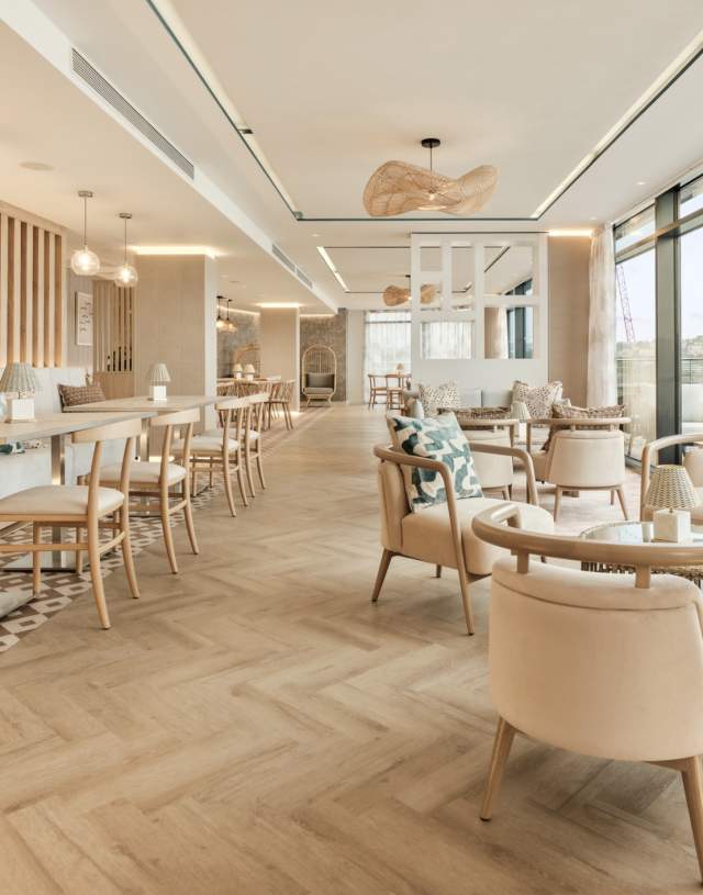Spacious modern café with light wooden floors, cosy beige seating, and large windows offering a scenic view. Warm, inviting atmosphere with stylish decor.