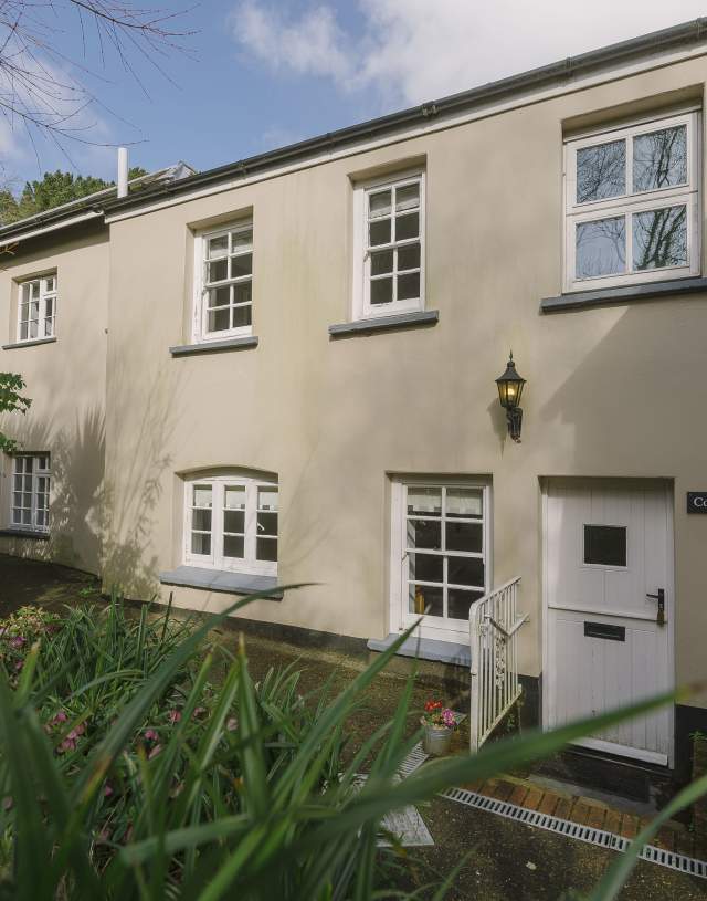 Two-story beige cottage with white-framed windows and a door, labeled "Corner Cottage." Surrounded by lush greenery under a partly cloudy sky.